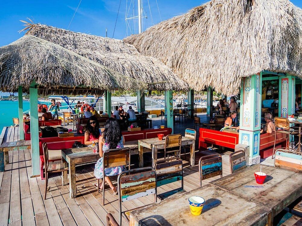 Daytime view of Bugaloe Beach Bar and Grill in Aruba showing an open-air over-water terrace with thatched palapa roofs, colorful painted pillars, rustic wooden tables and benches, guests dining, and boats moored at the dock visible in the background.