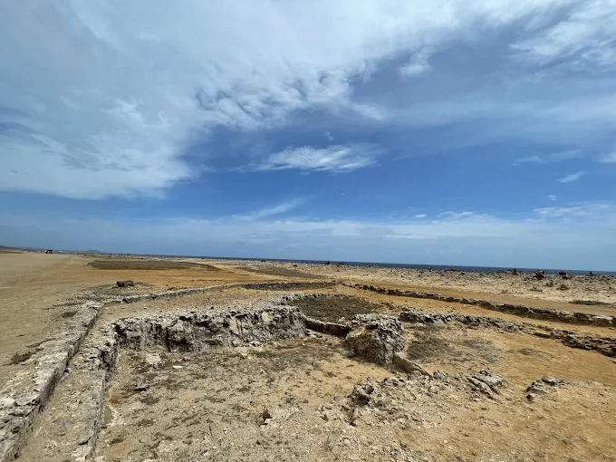 Wide view of ancient stone foundations along the sandy, arid coastline in Aruba under a partly cloudy sky.
