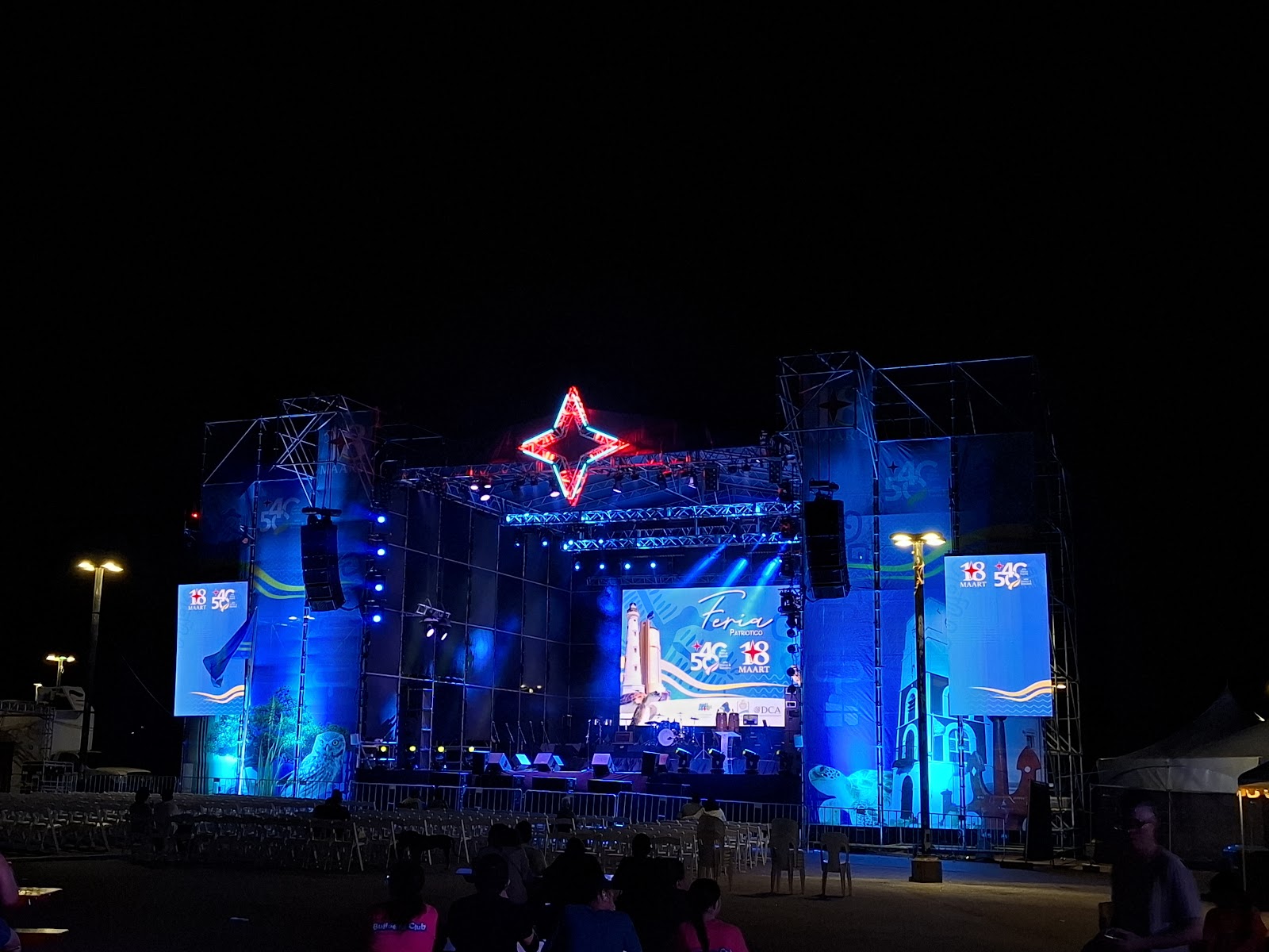 A large outdoor event stage illuminated with blue lighting, screens, and a star-shaped structure at night in Aruba.