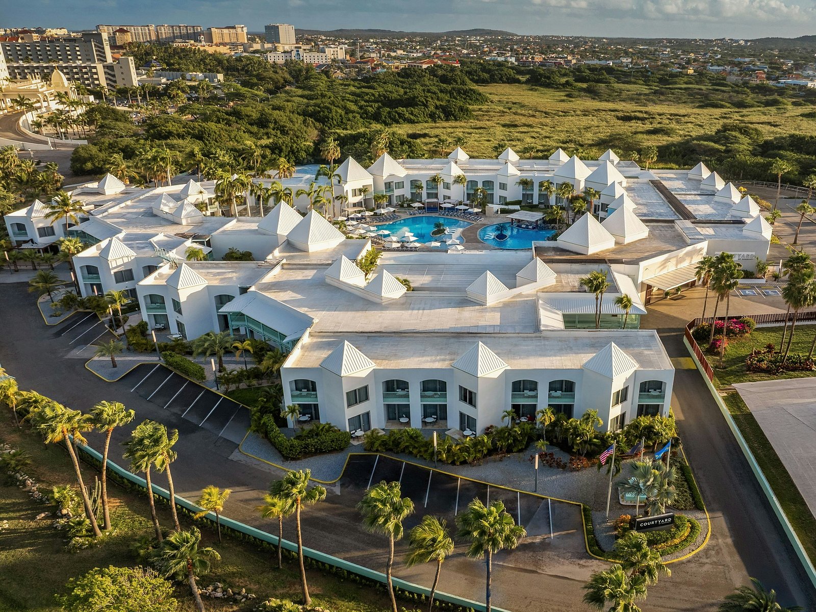 Aerial view of Courtyard by Marriott Aruba showing multiple buildings, the central pool area, parking, and surrounding landscape.