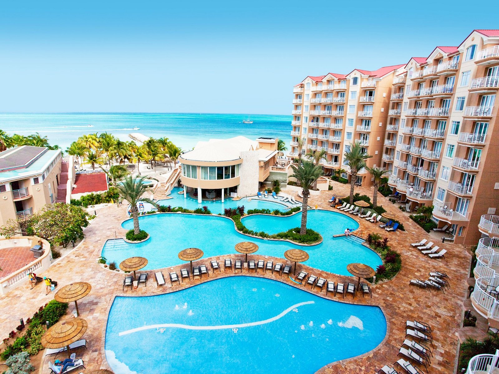 A free-form outdoor swimming pool with a wooden arch bridge, surrounded by palm trees, sun loungers, and resort buildings under a clear blue sky.