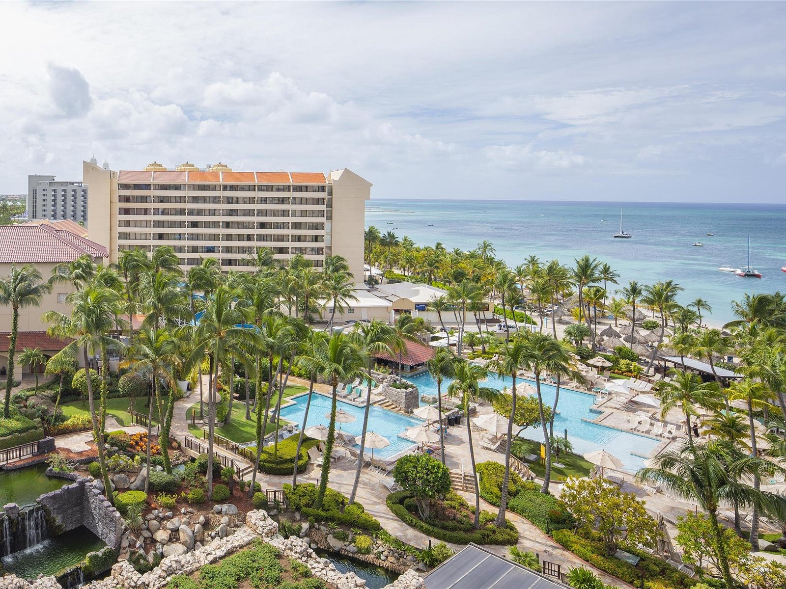 An elevated viewpoint overlooking winding resort swimming pools, dense palm trees, a multi-story hotel building, and the blue ocean coastline in Aruba.
