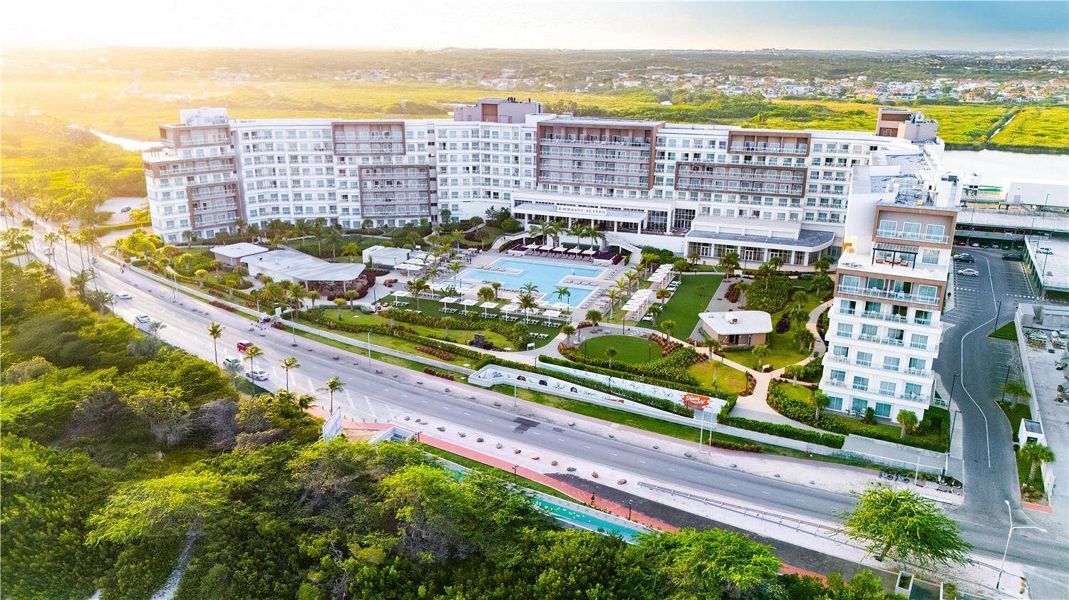 Aerial view of Embassy Suites by Hilton Aruba Resort showing the multi-story hotel building, outdoor pools, landscaped grounds, surrounding roads, and nearby greenery.