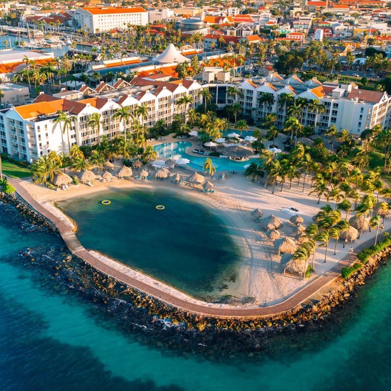Aerial view of Renaissance Ocean Suites in Aruba with resort buildings, a curved sandy beach, palm trees, swimming pool, and surrounding blue water.