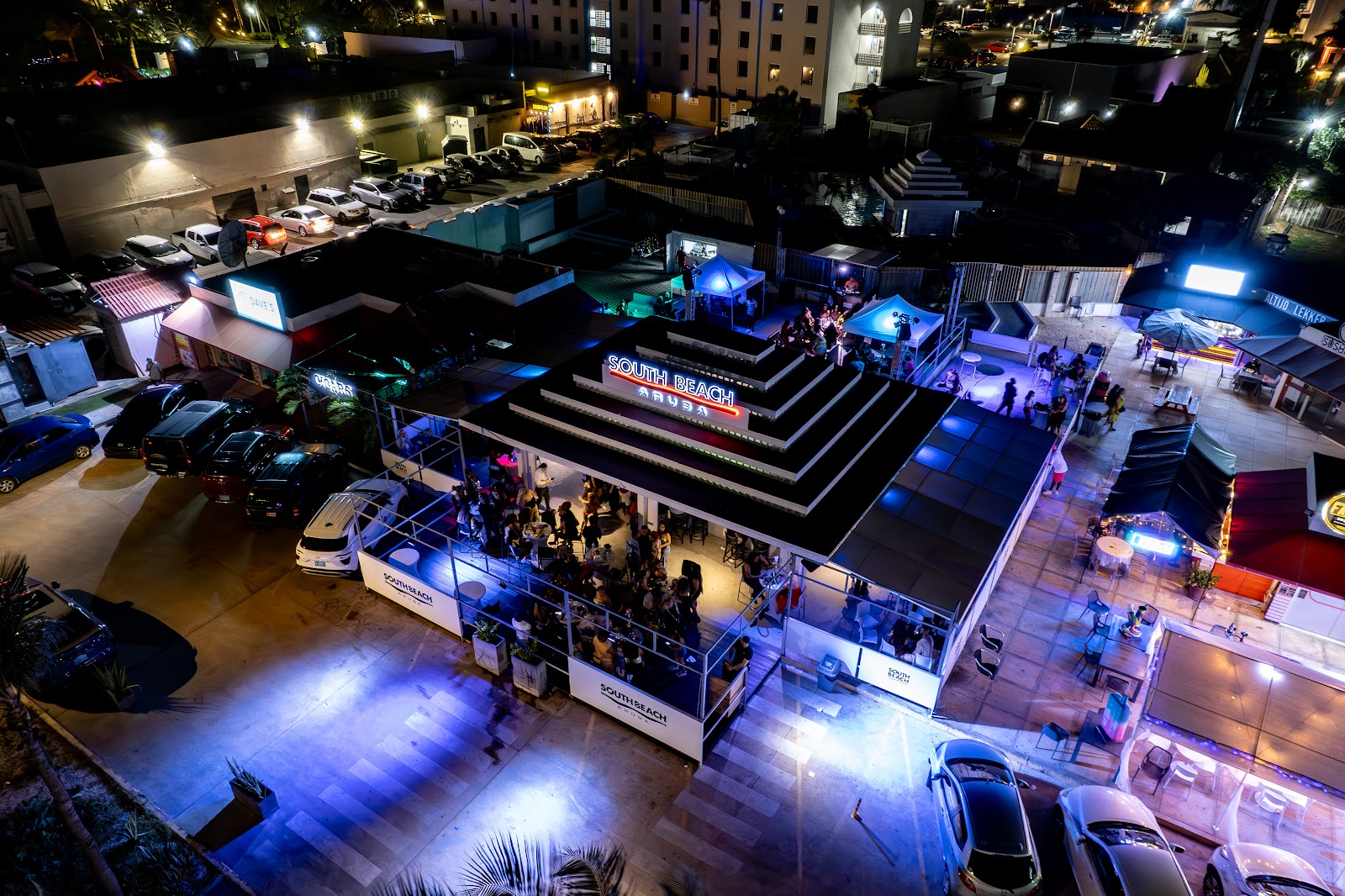 Aerial nighttime view of South Beach Aruba showing the restaurant and bar with illuminated outdoor seating and surrounding buildings.