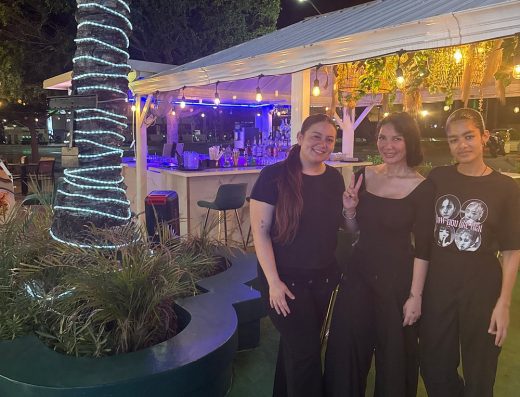 Three people posing beside the outdoor bar and illuminated palm tree at Chef Life Aruba Restaurant at night.
