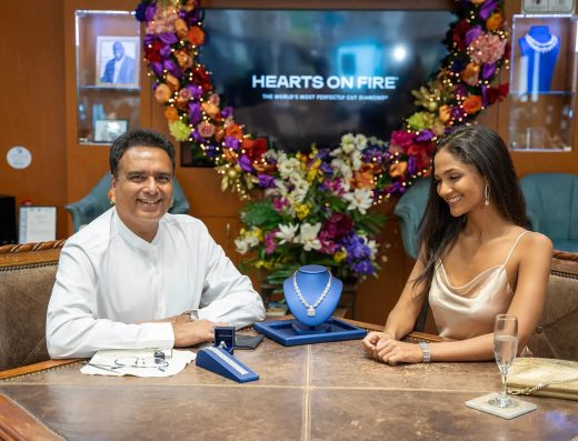 Two people seated at a jewelry counter inside Noble Jewelers in Aruba with a necklace display, jewelry pieces, flowers, and Hearts On Fire signage behind them.