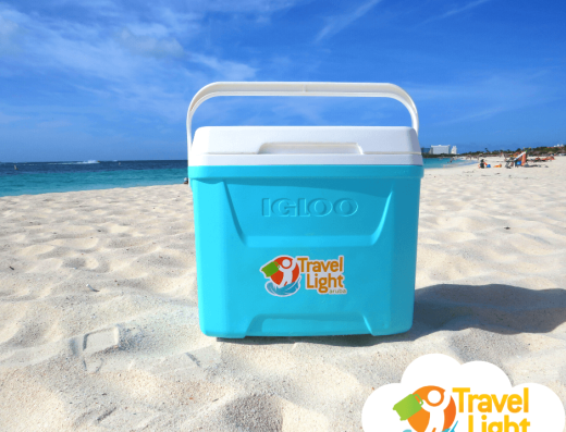 Blue Igloo cooler from Travel Light Aruba sitting on a white sandy beach beside turquoise Caribbean water.