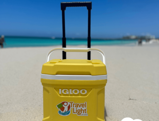 Yellow rolling Igloo cooler from Travel Light Aruba on a sandy beach with turquoise Caribbean water in the background.